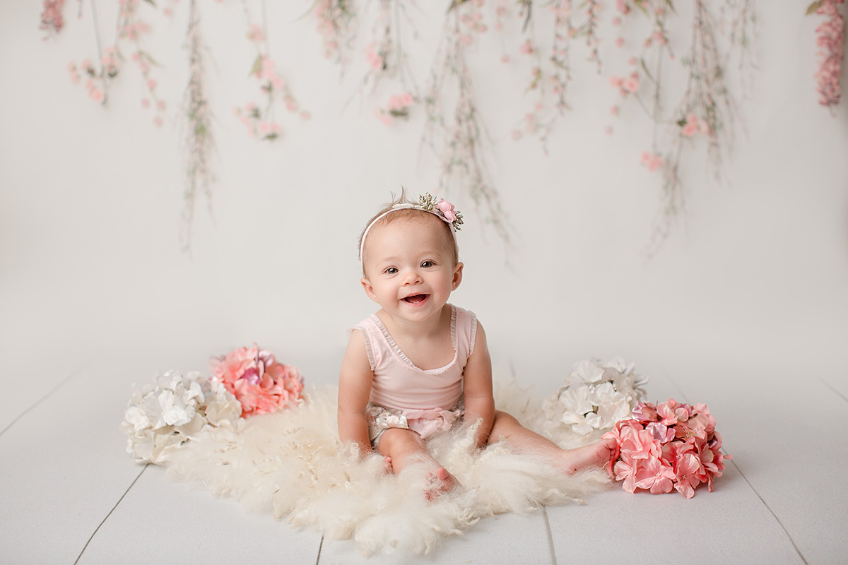 A smiling baby surrounded by flowers sits in a pink outfit and floral headband