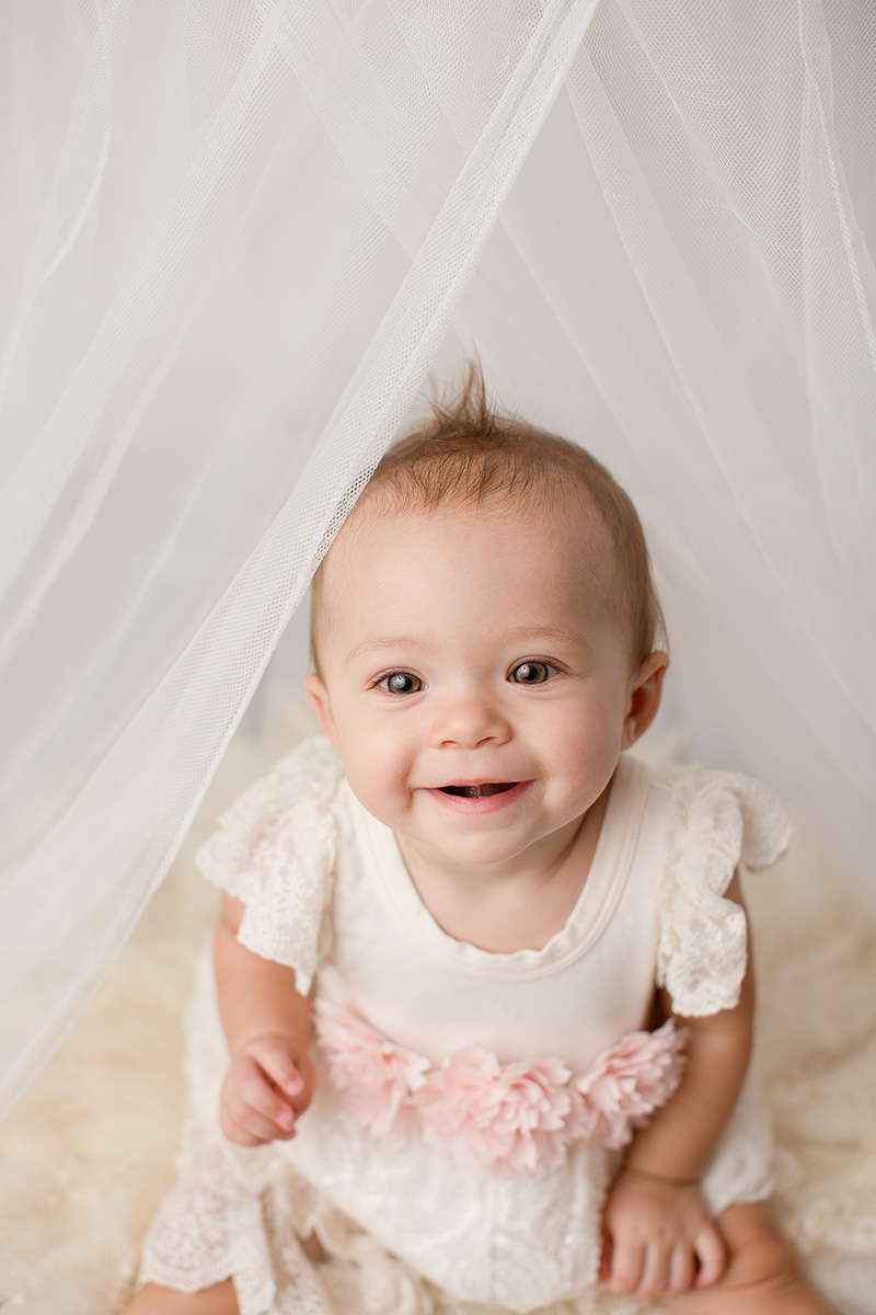 A happy infant in a white dress sits smiling under a curtain