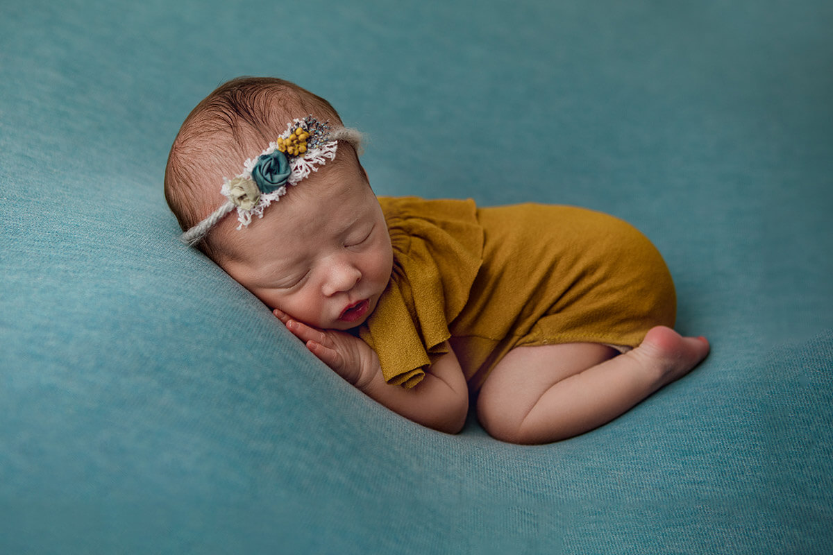 A baby girl sleeps in froggy pose in a brown dress and floral headband