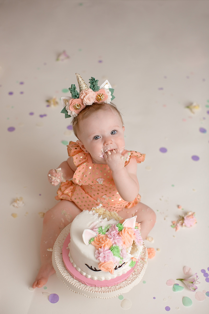 A happy baby looks up while shoving cake in her mouth in her orange dress and floral unicorn headband