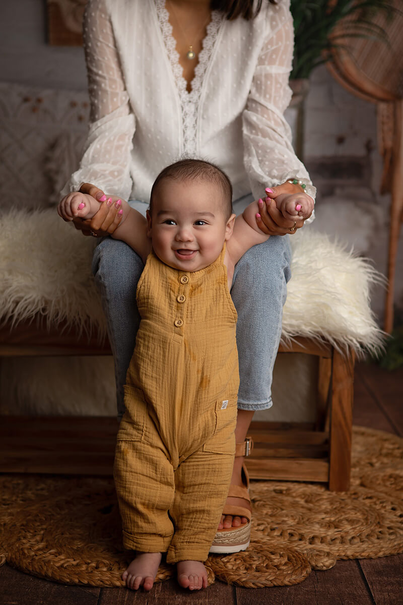 A happy baby in brown overalls stands with mom's help in a bedroom with a big smile after finding baby clothes in springfield il