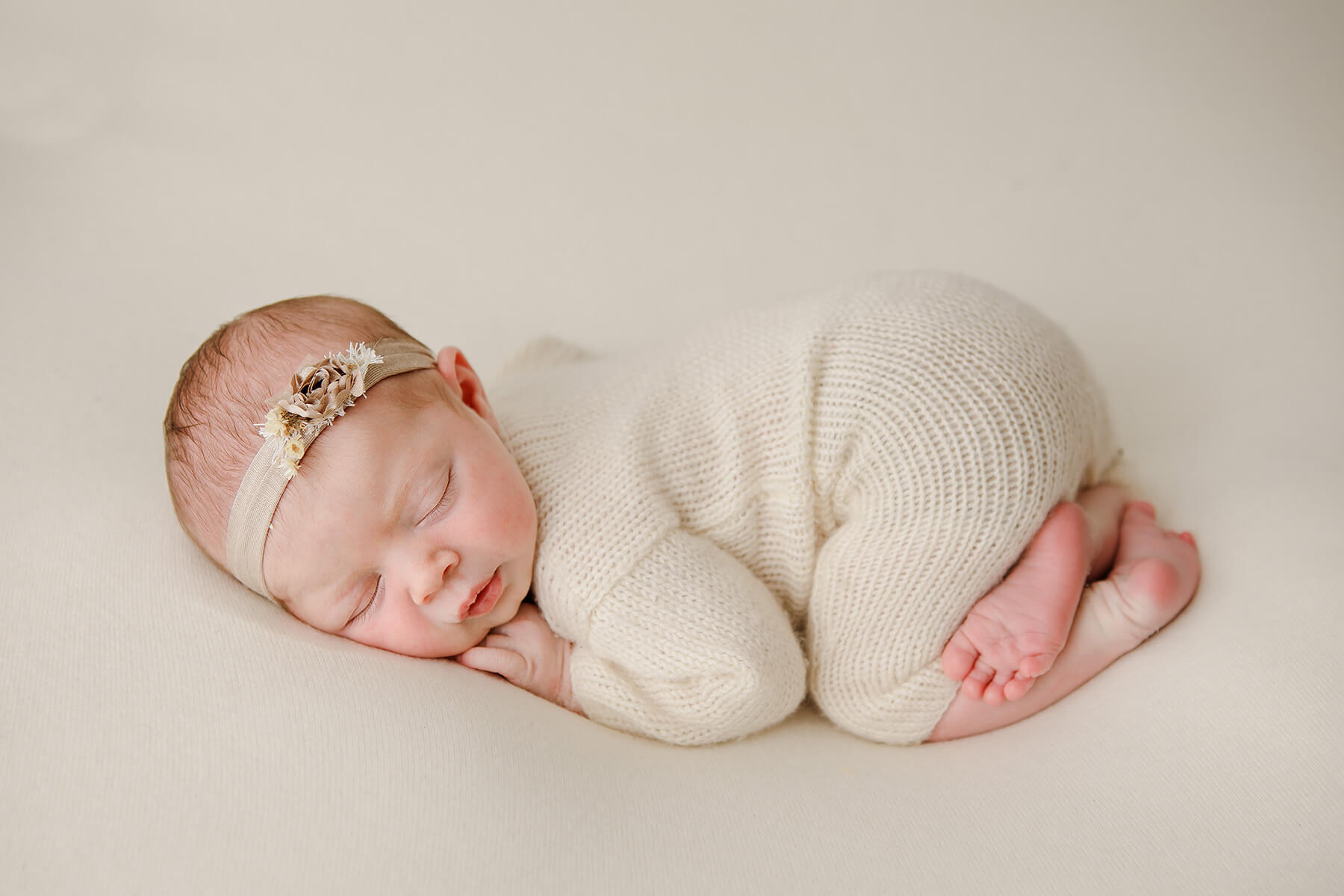 A newborn girl in a white knit onesie sleeps in froggy pose with a floral head band after some baby swim lessons in springfield il