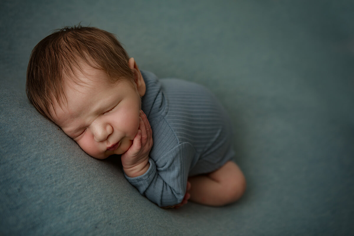 A baby boy in a blue onesie sleeps in froggy pose with a hand on his cheek in a studio after some baby swim lessons in springfield il