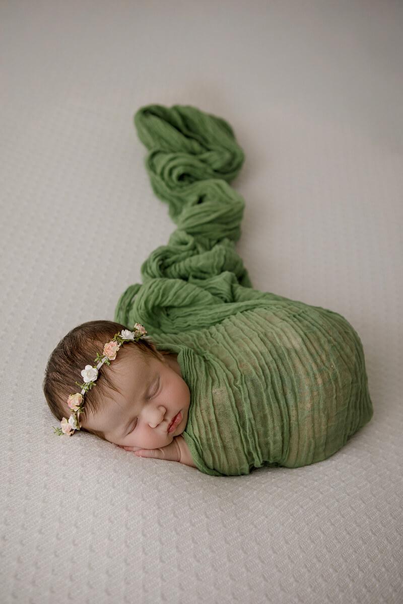 A sleeping newborn baby in a green swaddle and floral headband