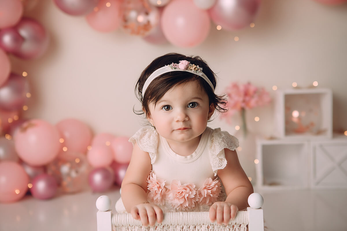An infant girl leans on the back of a white chair in a pink dress
