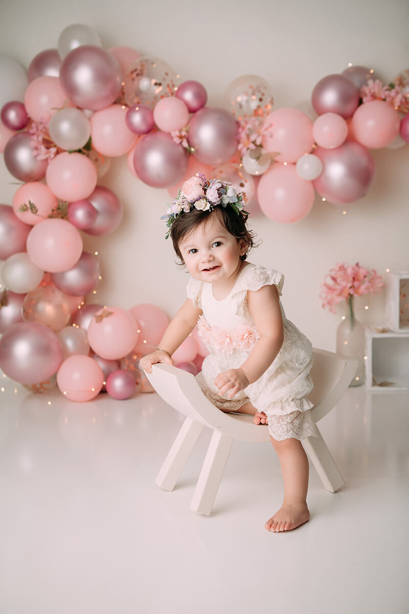 A happy infant girl plays on a curved wooden chair in a studio with a pink balloon wall after visiting daycares in springfield il