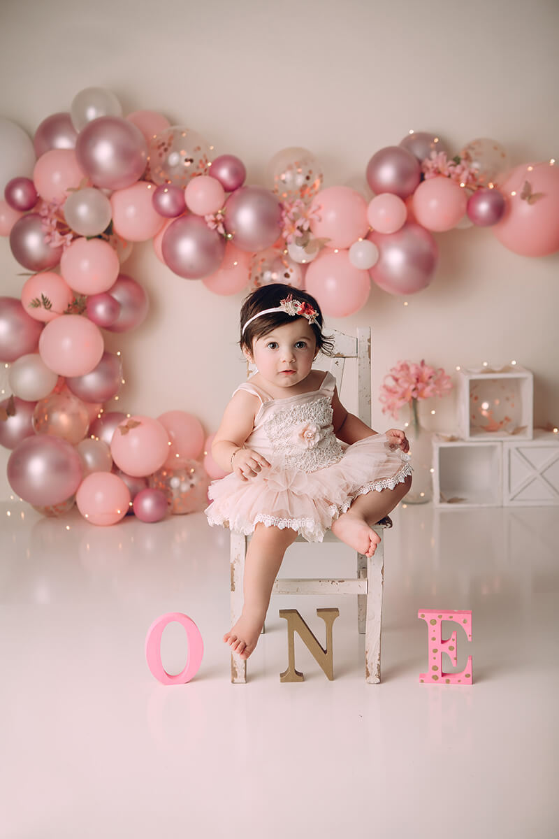 An infant girl sits on a small wooden chair for her first birthday with a pink balloon wall after visiting daycares in springfield il