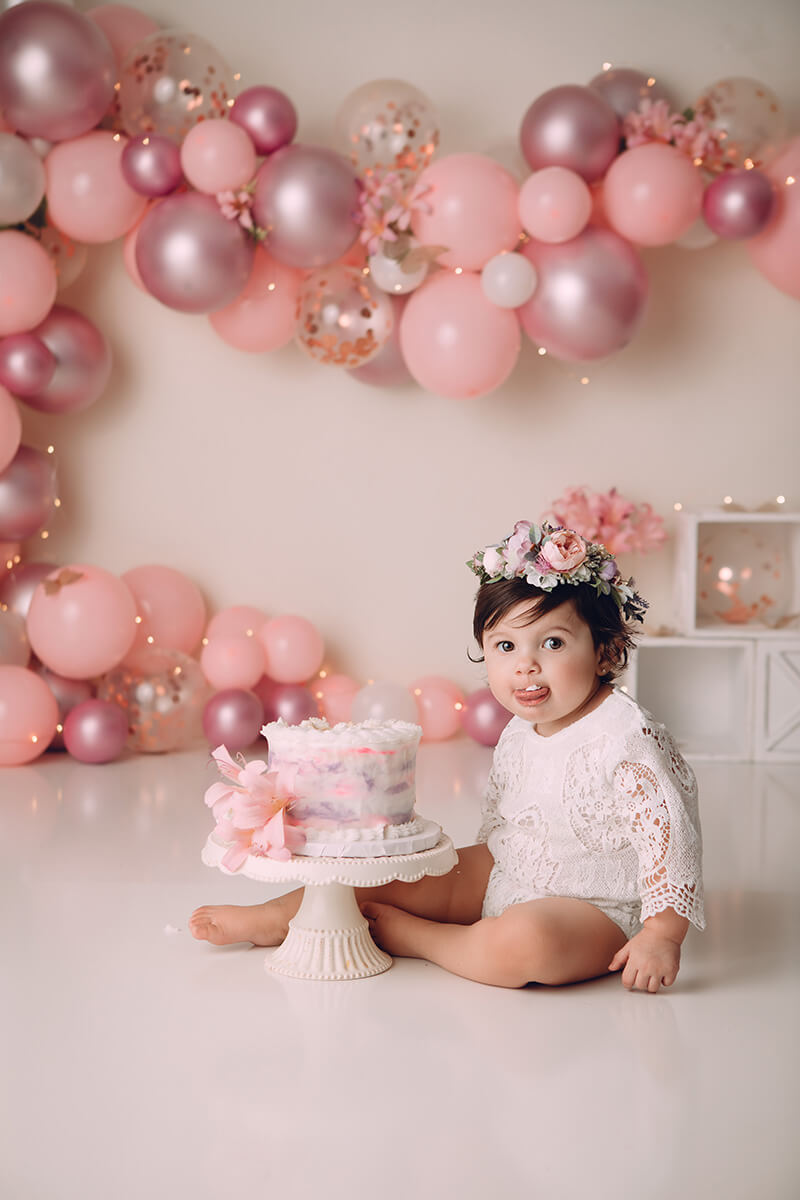 An infant in a lace shirt sits eating cake in a studio with a pink balloon wall after visiting daycares in springfield il