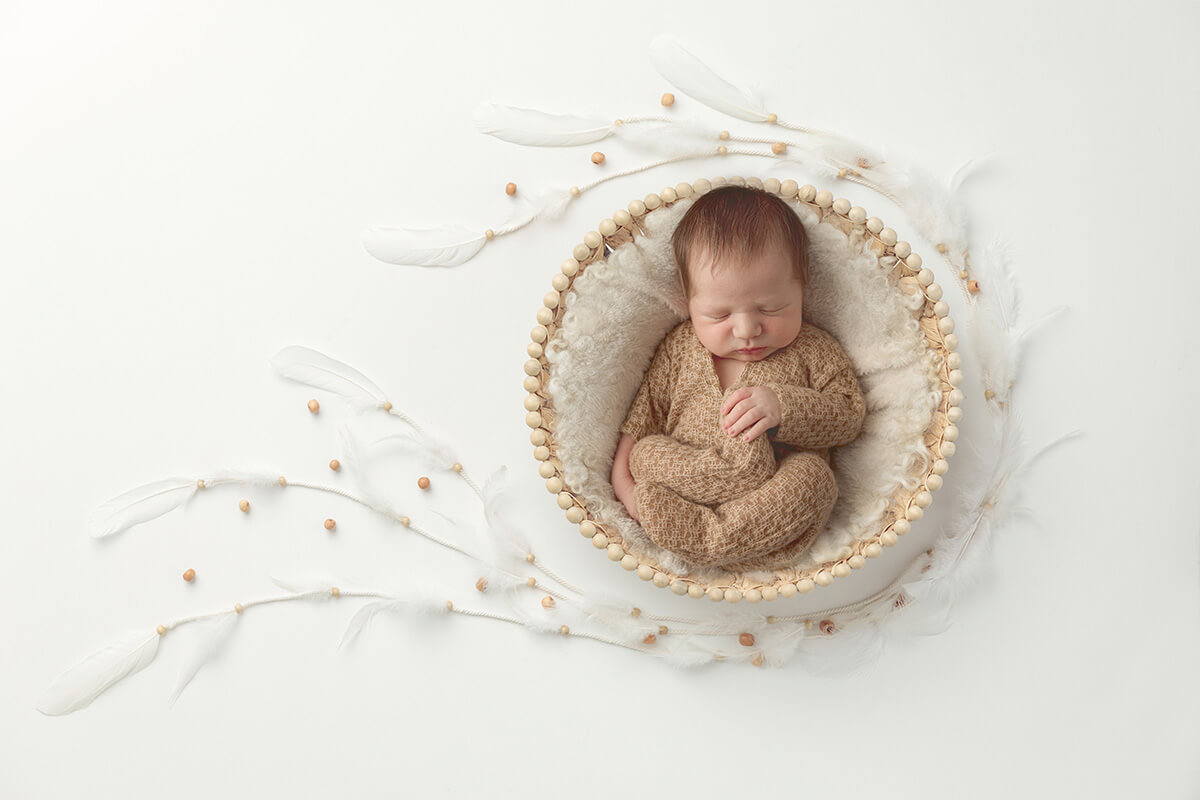 A baby sleeps in a brown onesie in a wooden bucket in a studio