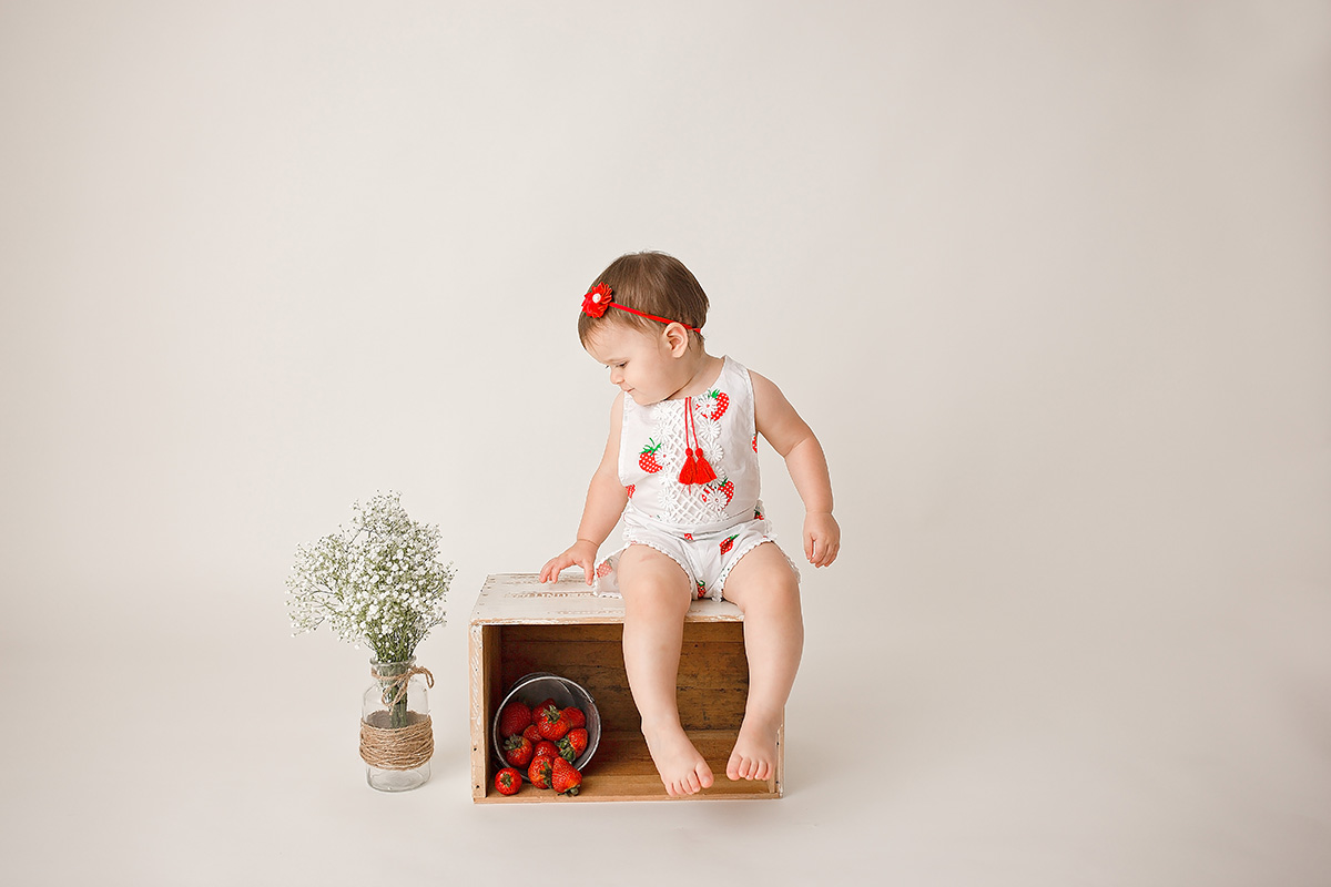 A smiling baby girl sits on a wooden box with a bucket of strawberries in a studio in a white strawberry onesie