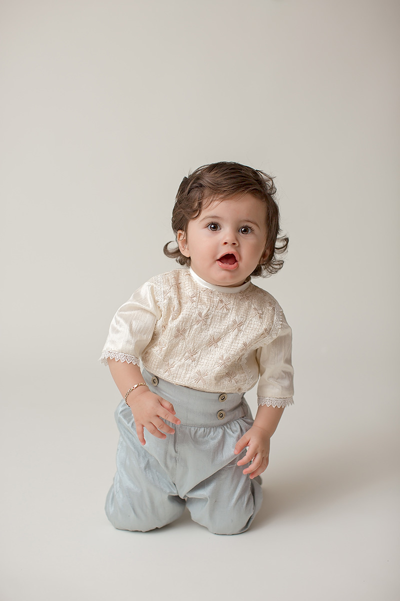 A happy toddler in blue pants and shiny white shirt smiles while kneeling in a studio