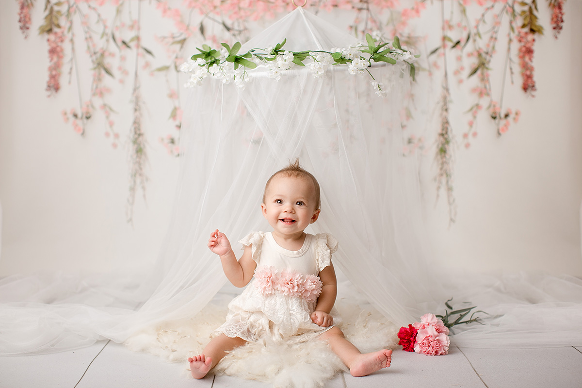 A happy toddler in a white dress sits under a tule canopy with flowers in a studio before visiting indoor playgrounds in springfield il