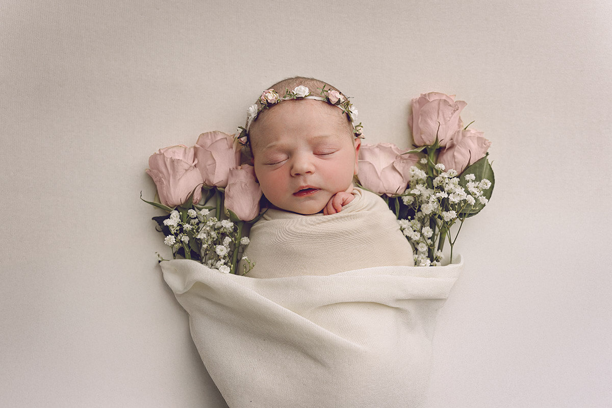 A newborn baby sleeps in a white swaddle surrounded by pink roses after meeting lactation consultants in springfield il
