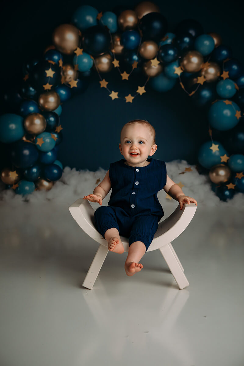 A happy baby in a blue onesie sits on a curved bench chair in a studio