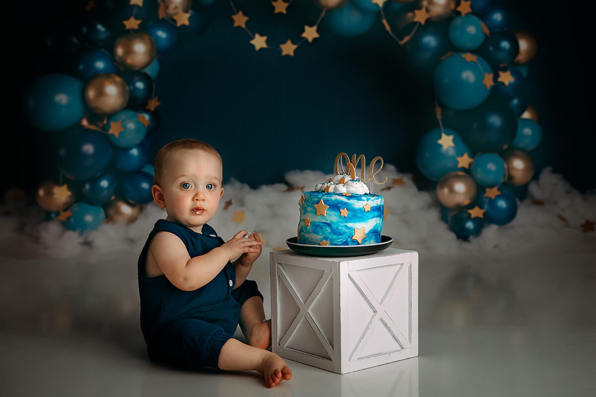 A baby sits with a birthday cake in a blue onesie on a wooden box after visiting little lincoln's toy shop