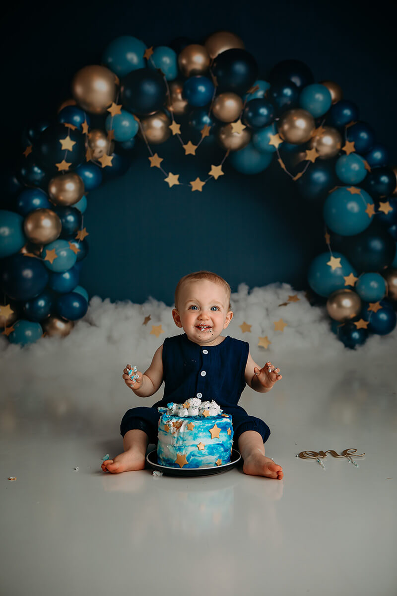 A happy baby in a blue onesie sits smashing his birthday cake after visiting little lincoln's toy shop
