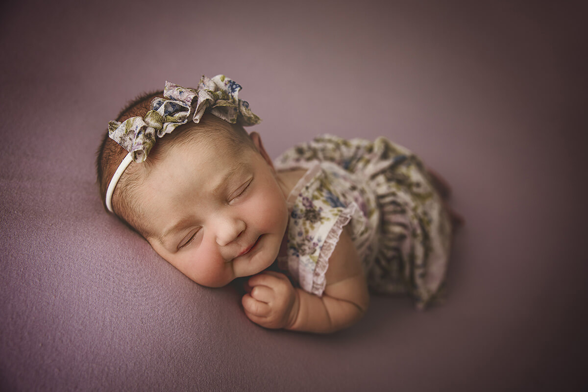A baby girl smiles in her sleep in froggy pose ina. floral dress and matching headband
