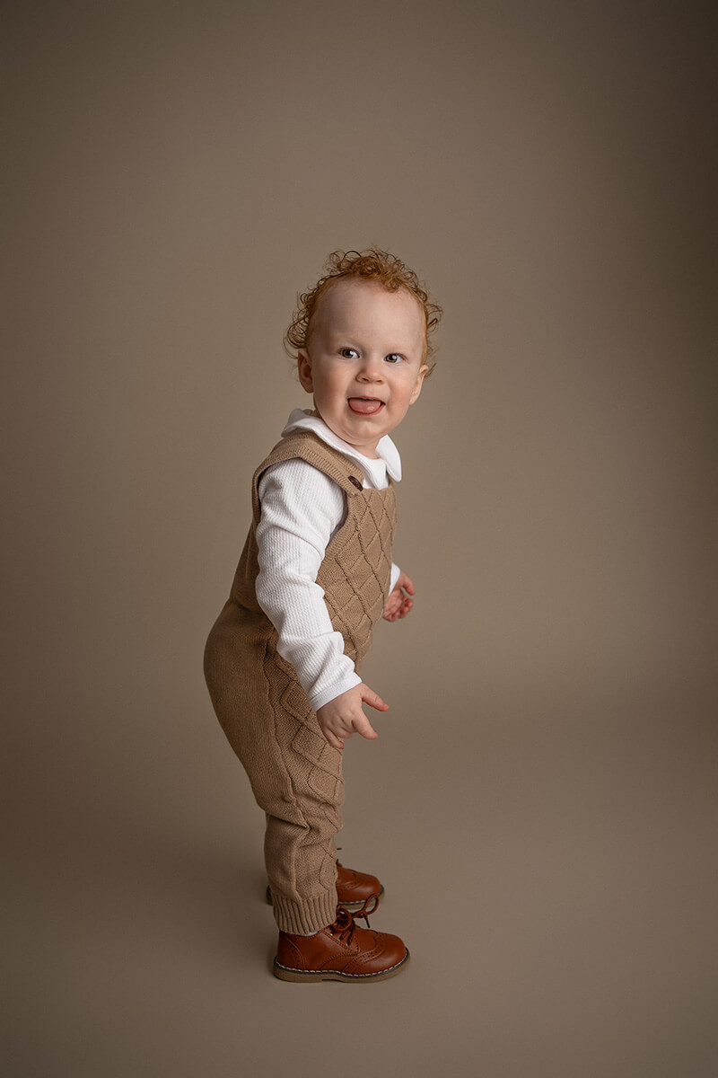 A happy baby in brown knit overalls stands with tongue out in a studio