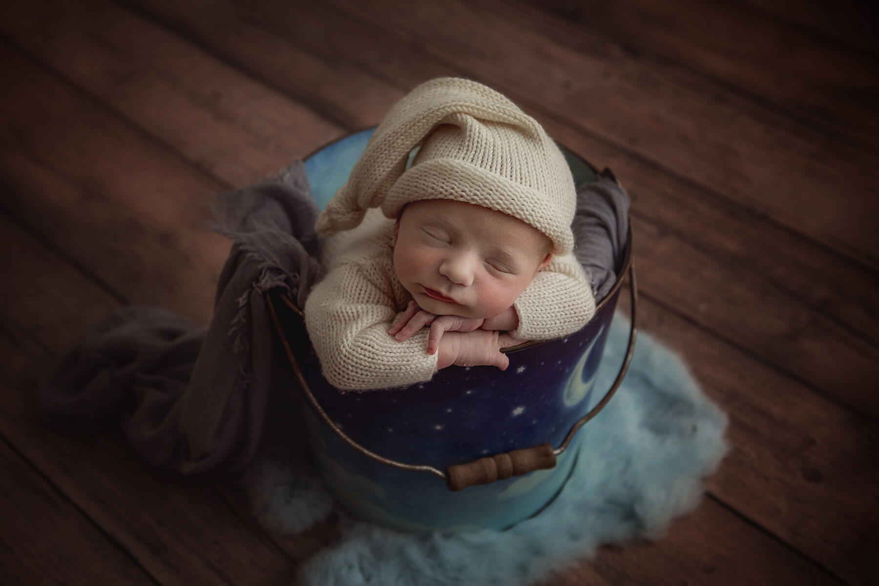 A newborn baby sleeps in a tin bucket in a white knit onesie and night cap