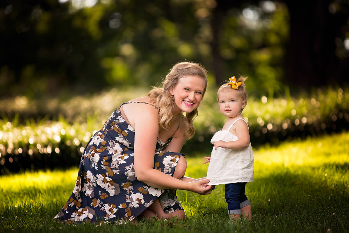 A smiling mom in a floral print dress kneels to help her toddler daughter fix her dress in a lawn at sunset after visiting playgrounds in springfield il