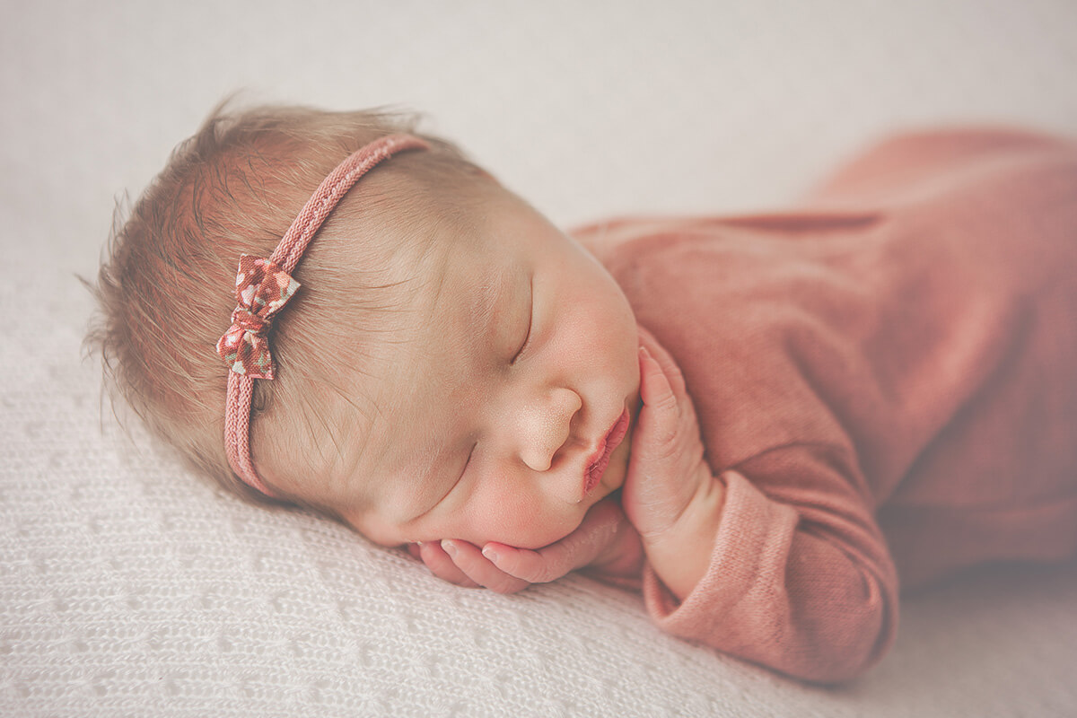 A newborn baby sleeps with hands on cheeks in a pink onesie