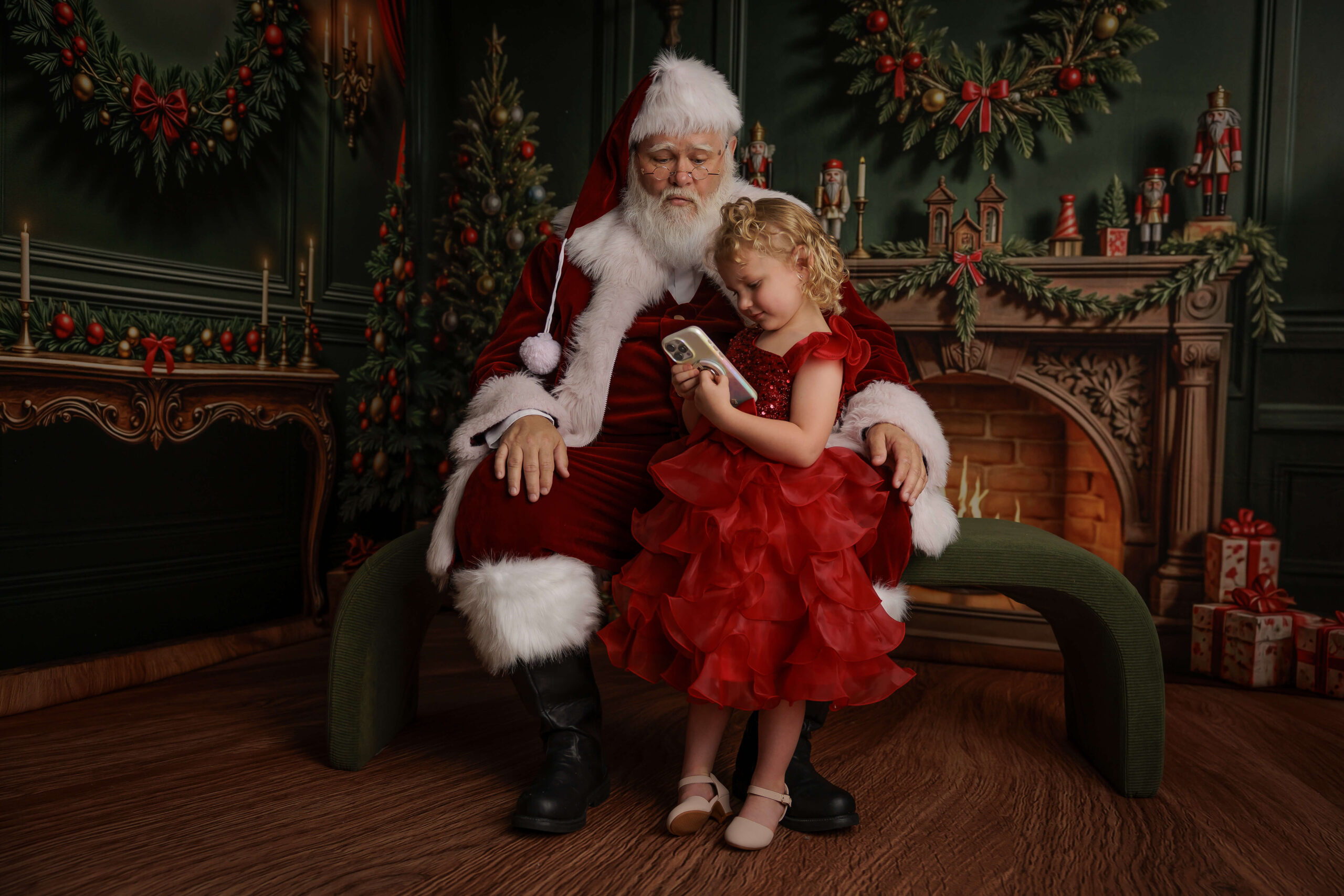 A young girl ina. red dress leans against a Santa in Springfield, IL sitting on a green bench in front of a fireplace