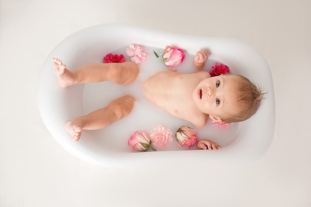 A baby lays in a murky bath tub with pink flowers floating in the water