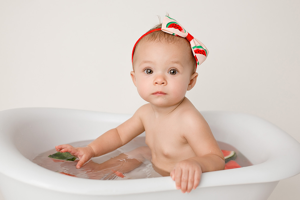 A baby girl with a watermelon headband plays with some watermelon slices in a bathtub after visiting splash pads in springfield il