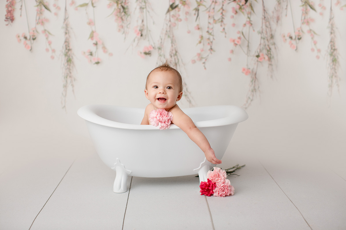 A smiling baby girl with pink carnations sits in a bath tub in a white studio with one arm hanging out