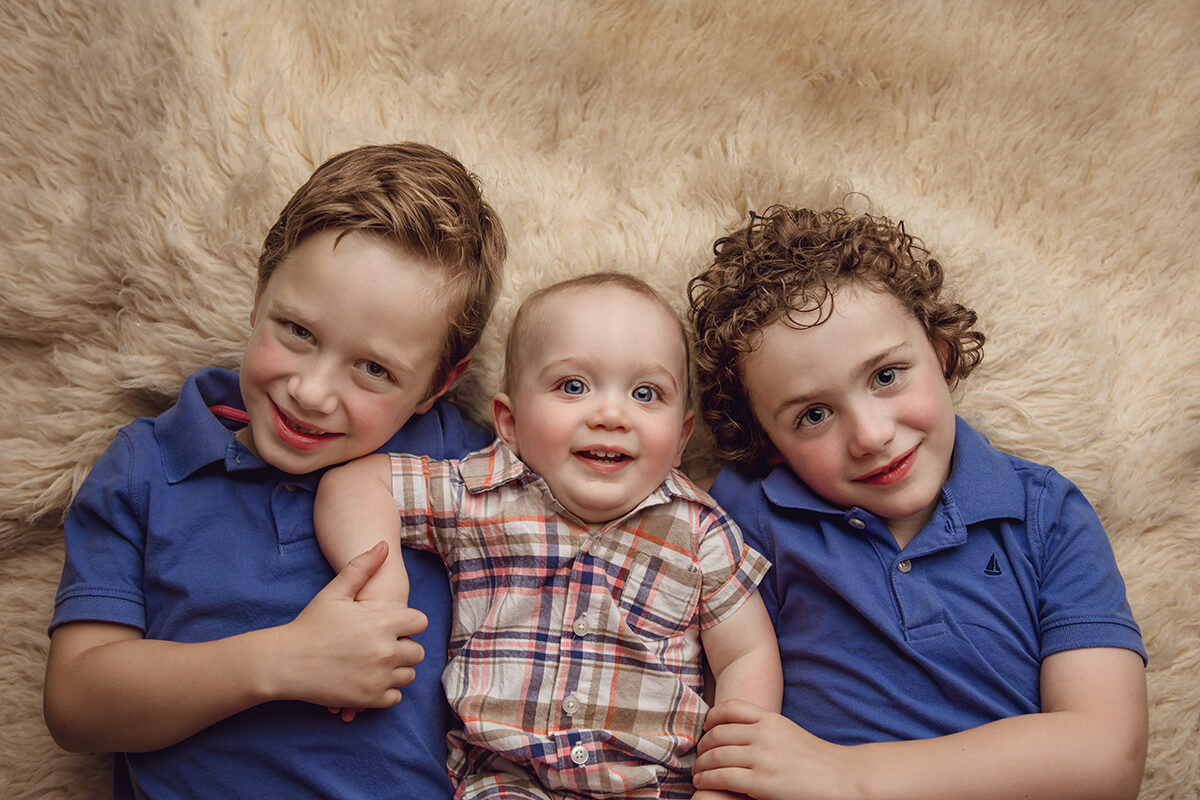 A happy baby in plaid lays surrounded by his two big brothers in blue polos on a fur blanket