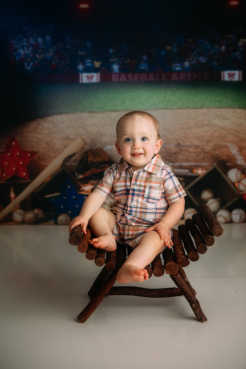 A toddler in a plaid shirt sits in a wooden log stool in a baseball themed studio smiling before some things to do in springfield il with toddlers