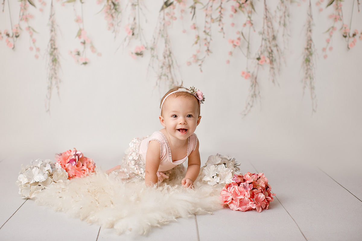 A baby girl crawls on a white fur blanket with a big smile in a pink dress