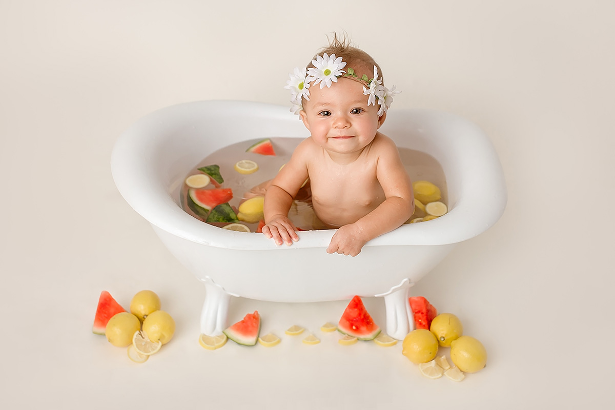 A smiling infant sits in a bath tub filled with water and fruit with a white flower headband in a studio after visiting toy stores in Springfield, IL