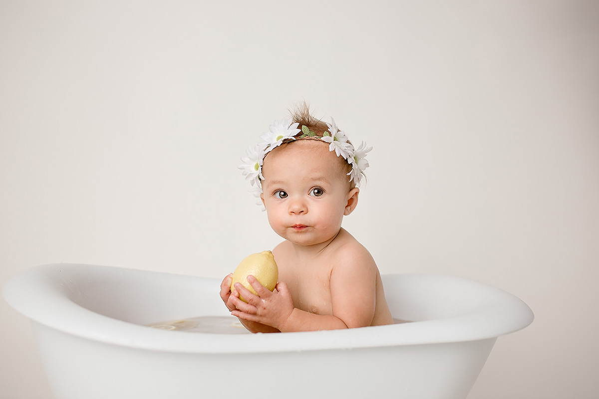 An infant holds a lemon and wears a white daisy headband while sitting in a bath tub