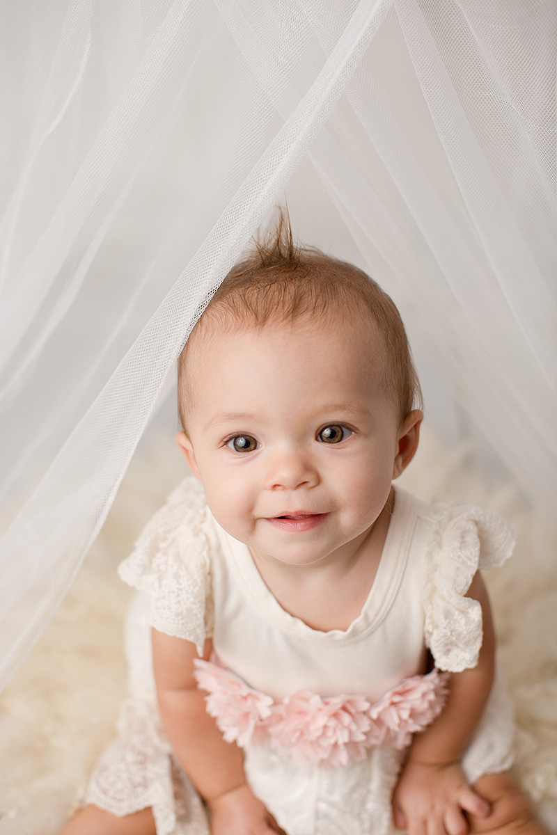 An infant girl in a white and pink dress sits smiling under some parted drapes before visiting toy stores in Springfield, IL