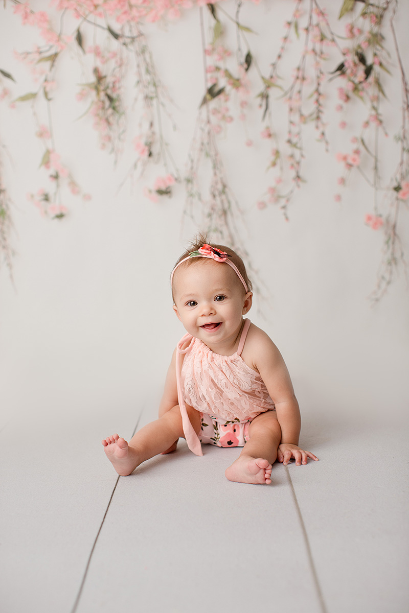 An infant girl in a pink top and matching floral headband sits on the floor of a studio smiling before visiting toy stores in Springfield, IL