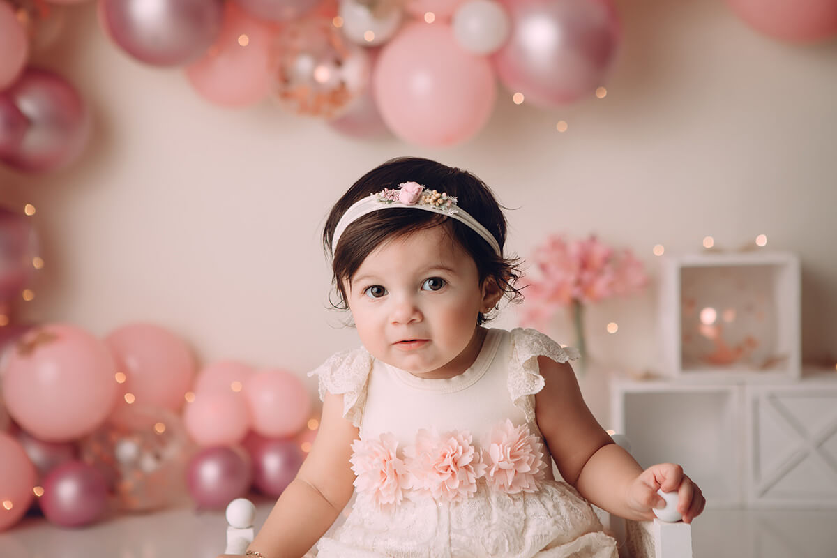 A young girl sits in a wooden chair in a pink dress with flowers in a studio