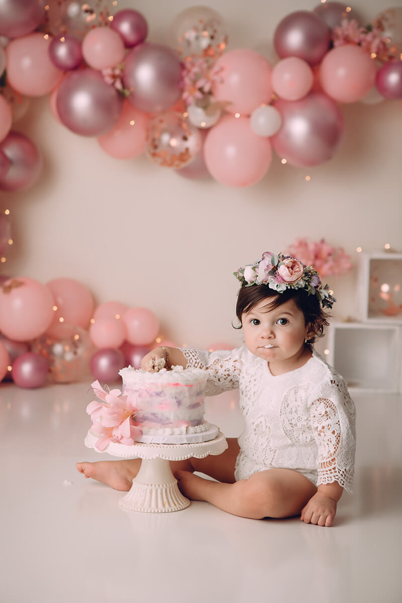 A baby girl in a lace white onesie sits eating cake in a studio with pink Balloons by Gabi on the wall