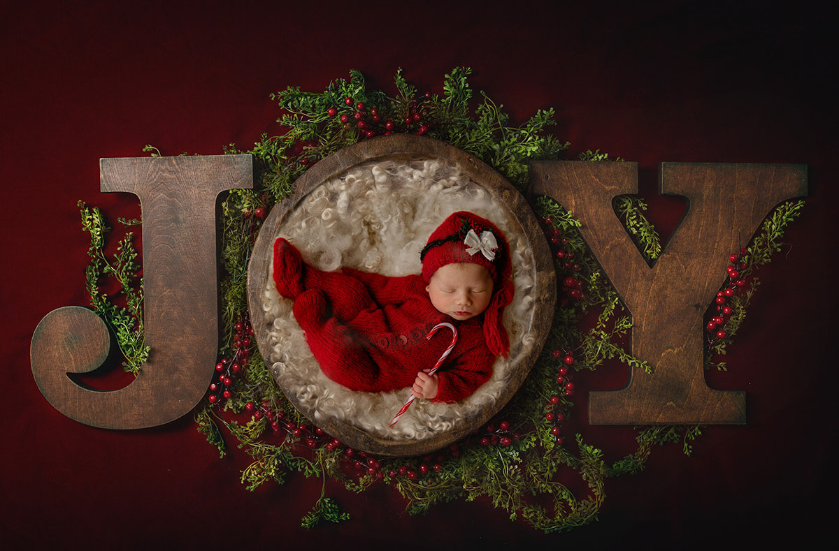 A sleeping christmas baby in a red knit onesie sleeping in the a wooden bowl spelling joy after leaving birthing centers in Springfield, IL