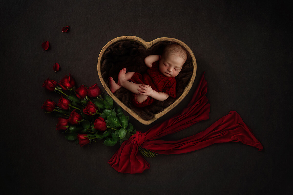 A sleeping newborn in a red knit onesie in a heart shaped bowl with red roses after leaving birthing centers in Springfield, IL