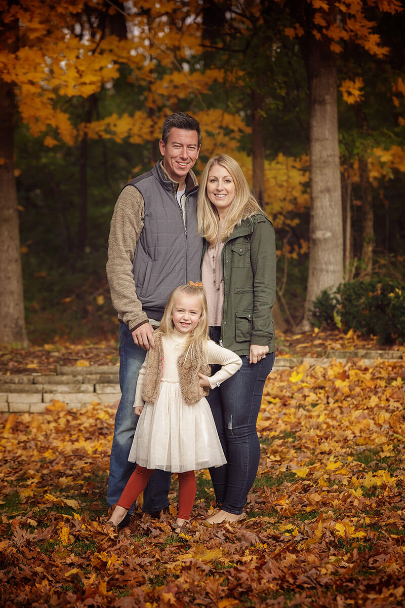 A mom and dad stand with their toddler daughter in a white dress in fall during some family activities in Springfield, IL