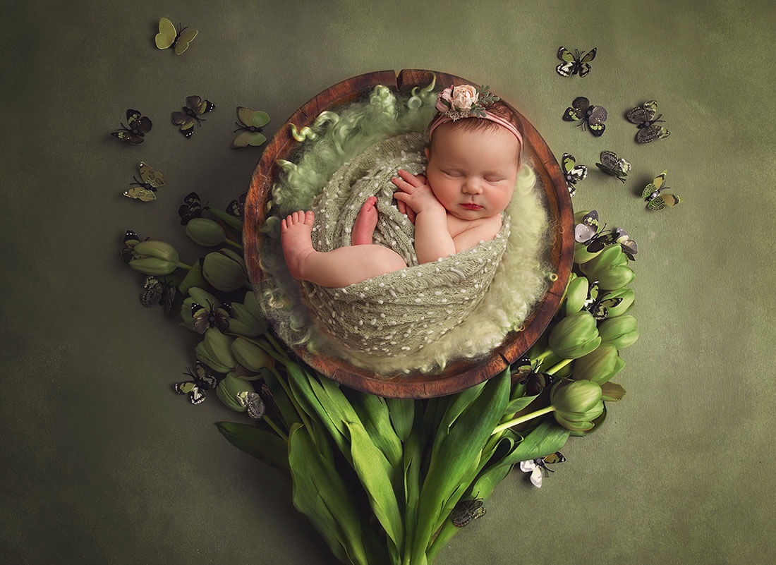 A newborn baby in a wooden bowl sleeping on a green blanket surrounded by tulips and butterflies