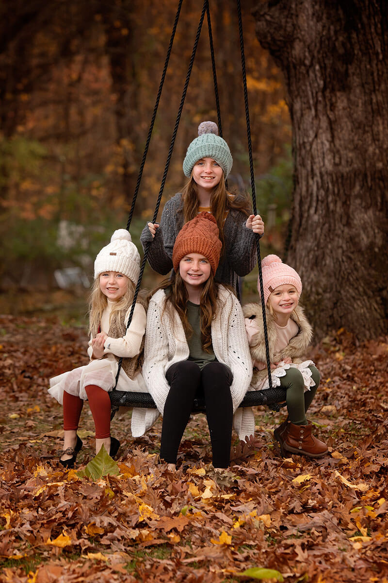 Four young cousins in sweaters and beanies sit on a tree swing smiling