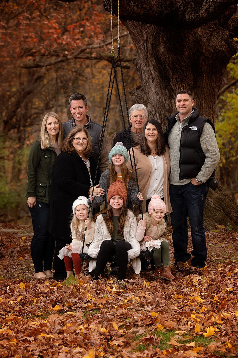 Four young cousins sit on a tree swing while smiliing with their extended family in fall in one of the parks in Springfield, IL