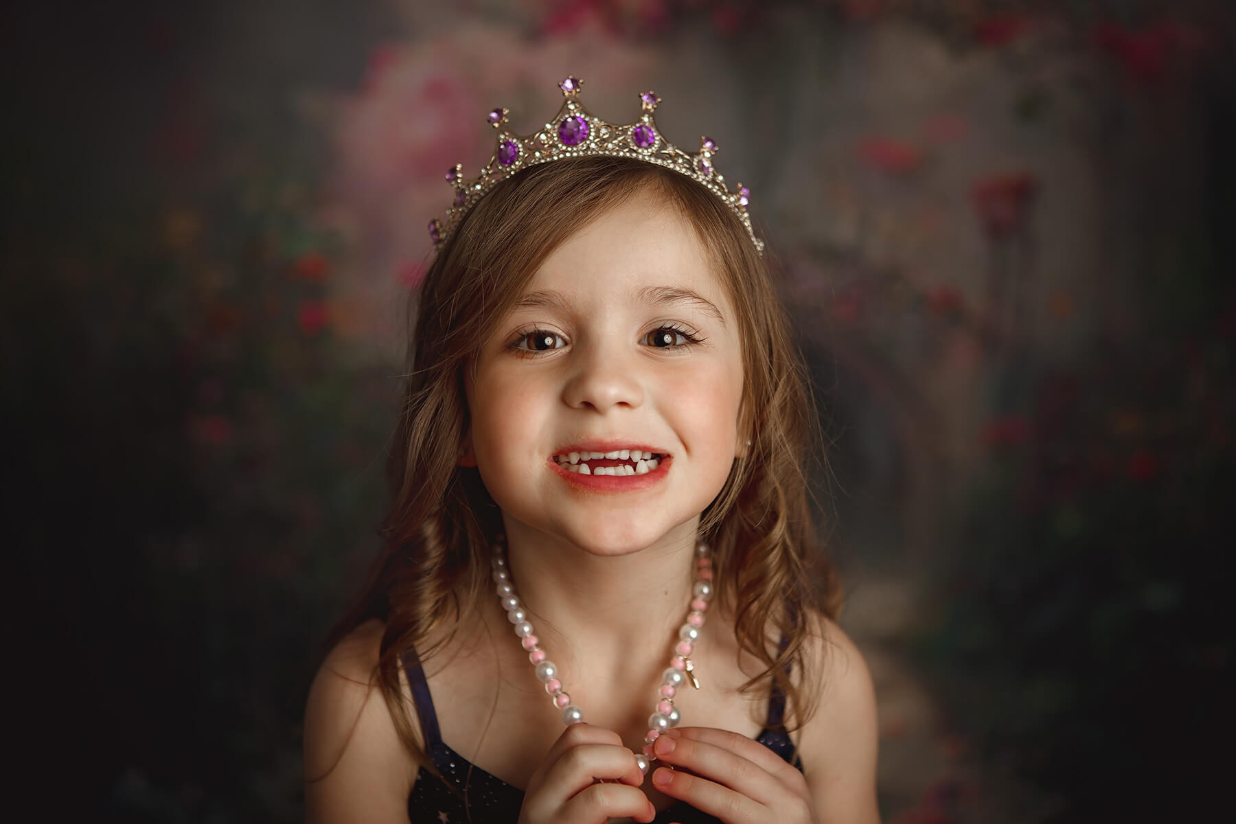 A closeup of a happy toddler girl with a tiara and pearl necklace in a studio