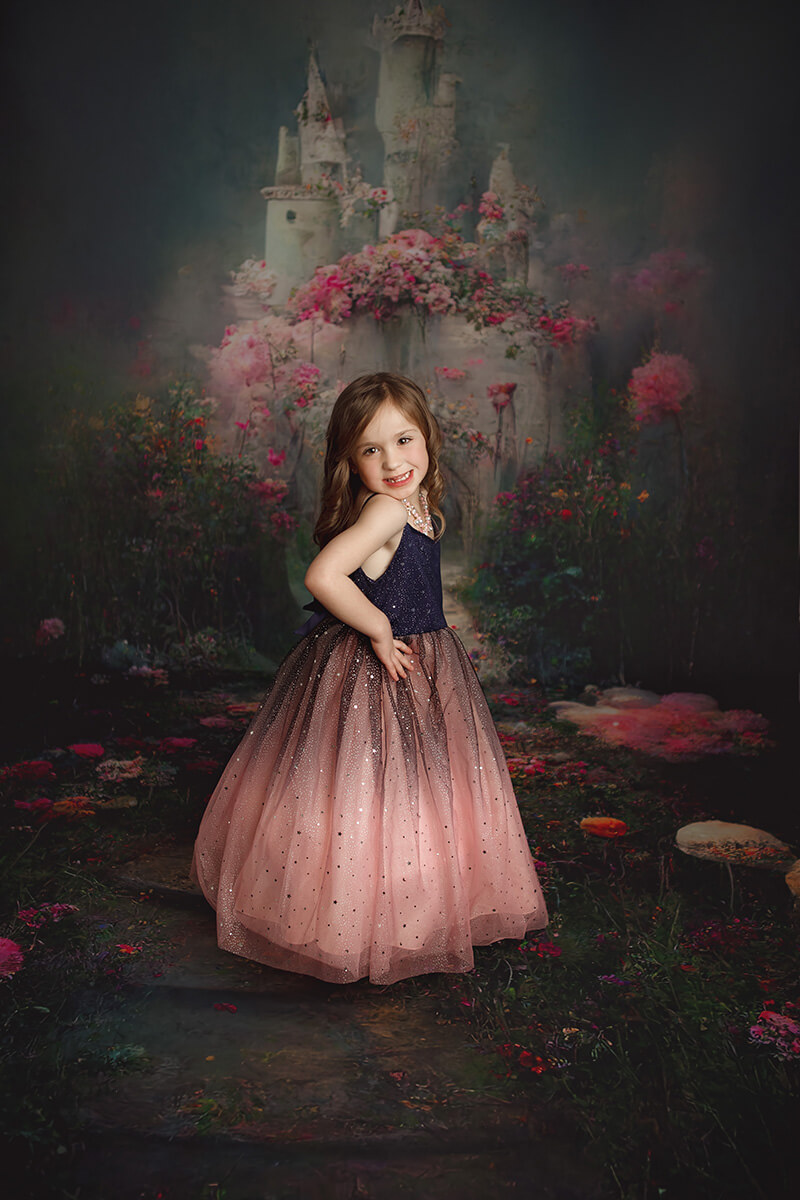 A smiling young girl with hands on her hips stands in a princess themed studio in pink and blue dress