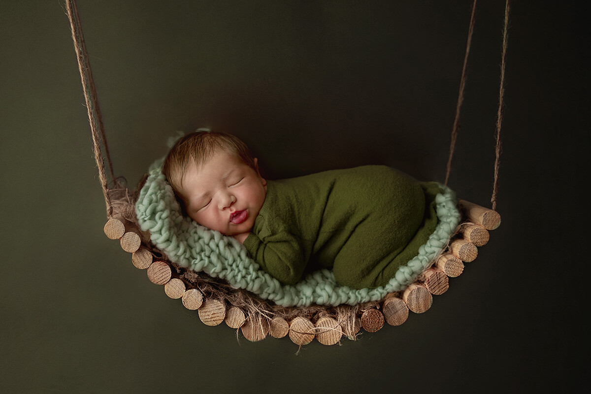 A sleeping newborn baby on a wooden swing and knit blanket in a forest green onesie in froggy pose