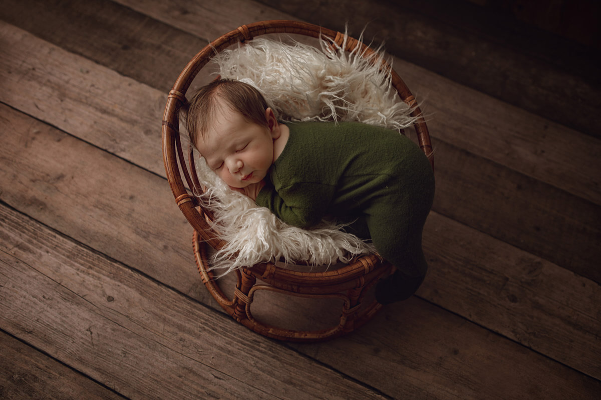 A newborn baby sleeps in a wicker chair on a fur blanket in a forest green onesie