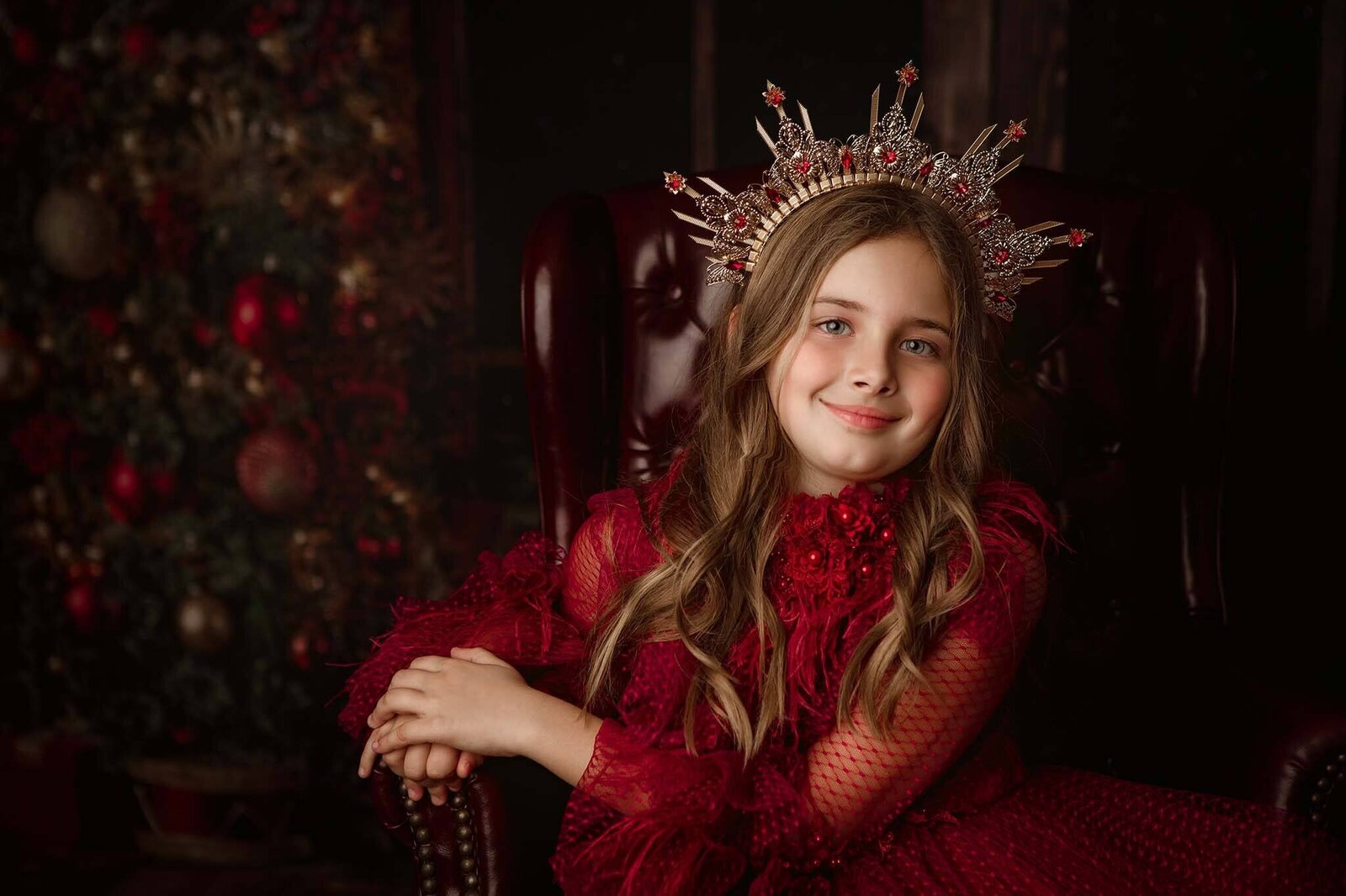 A happy young girl in a red gown sits on an antique chair with a large crown in front of a christmas tree