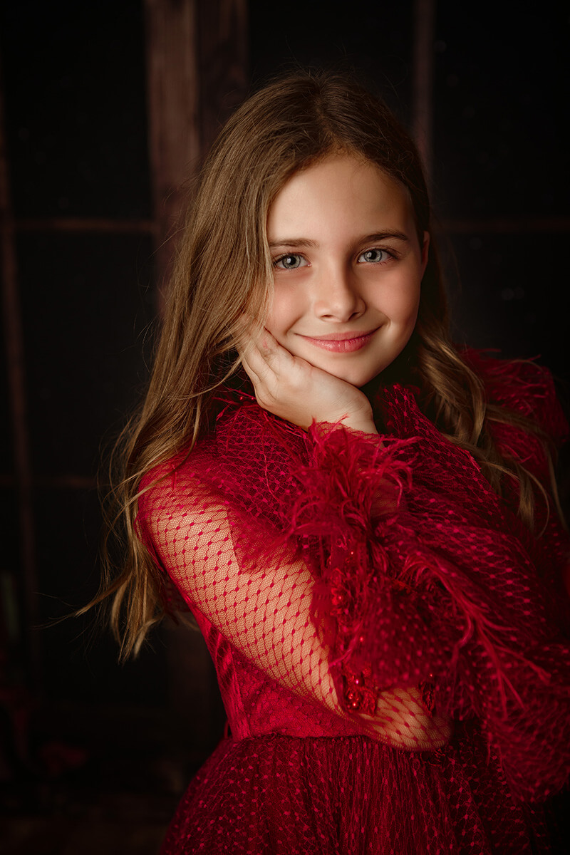 A young girl in a red gown rests her chin in her hand while smiling in a studio before visiting an apple orchard in Springfield, IL